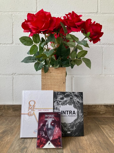 Fotografía de las tres novelas de Isabel Pedrero; a`pyadas sobre un florero de madera con varias rosas, Omega, Intra y 999 pedazos